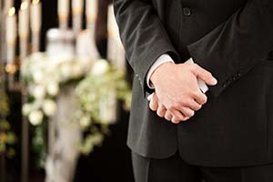 En person i svart kostym står med händerna över varandra. I bakgrunden vita sorgeblommor